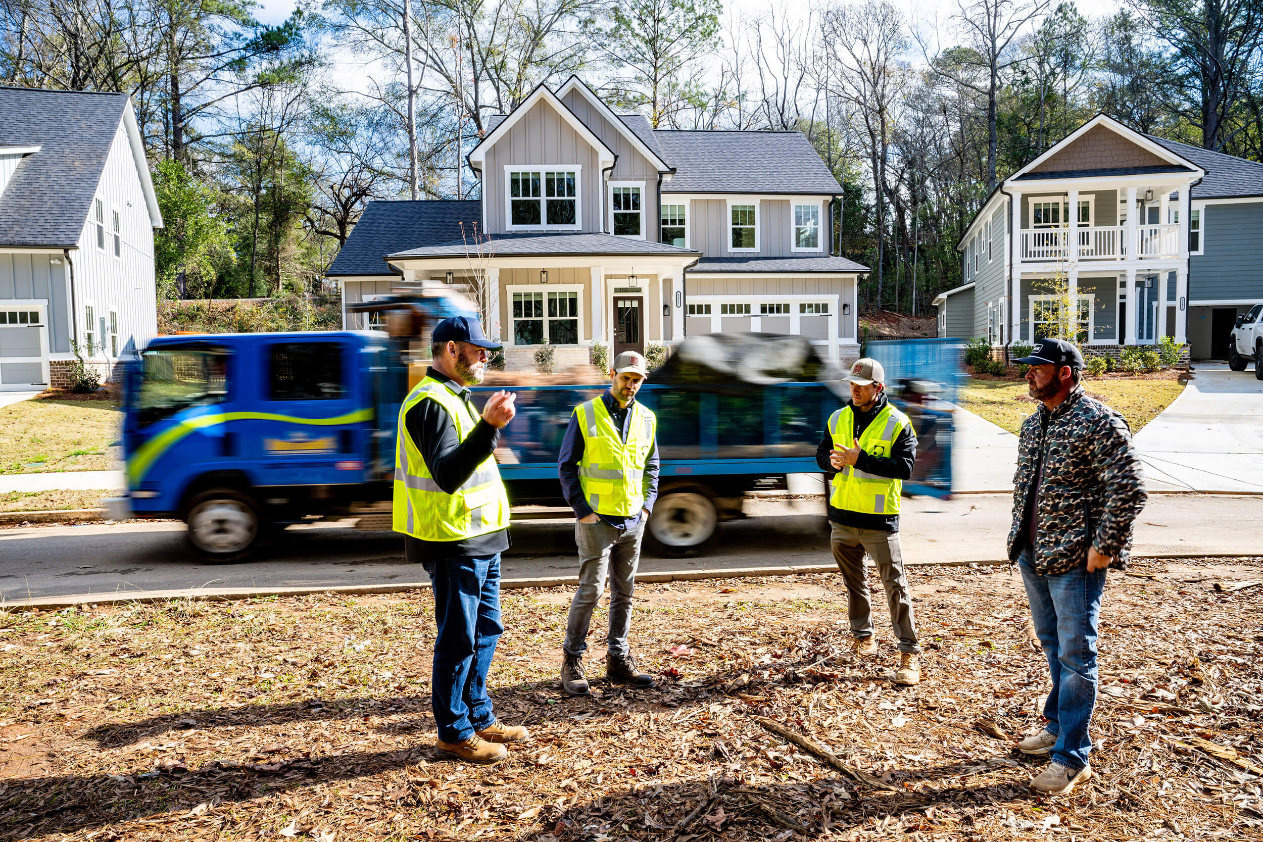 Four construction workers in high-visibility vests discuss in front of modern houses and a passing blue truck.