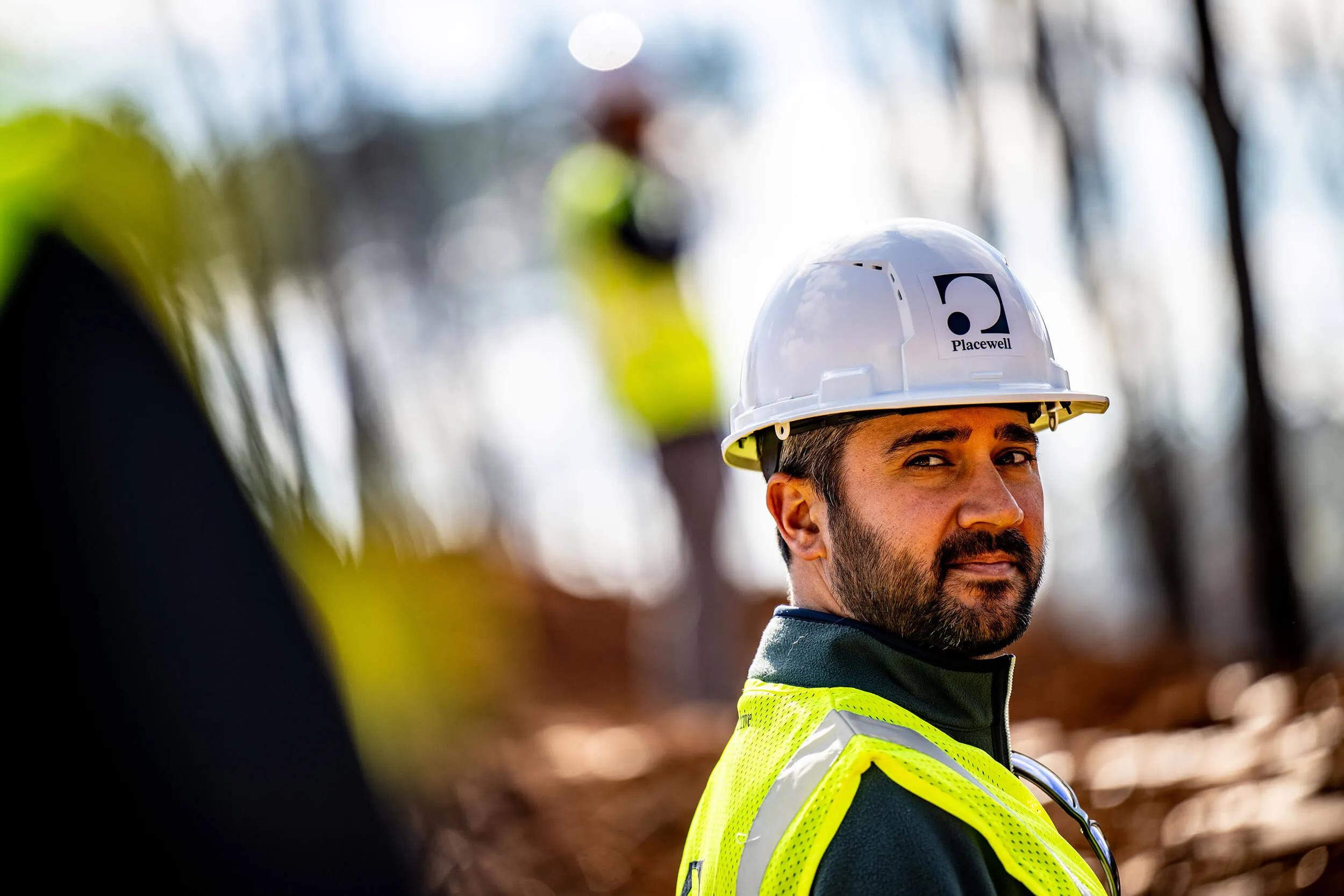 Worker in a white Placewell hard hat and neon safety vest looking at the camera outdoors.