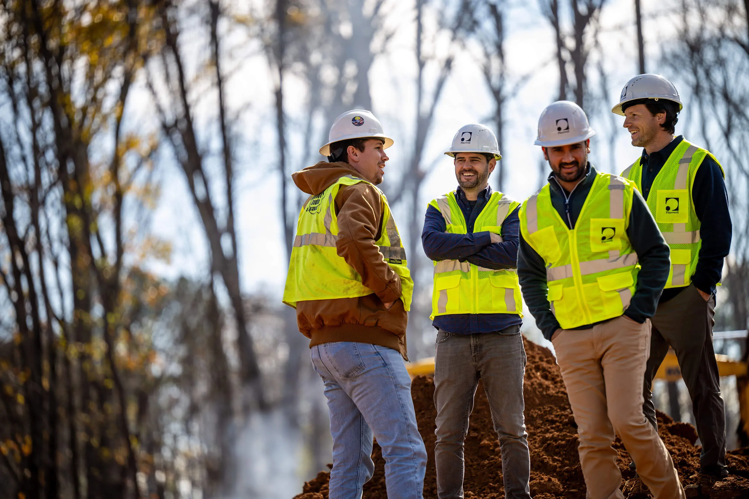 Four construction workers in helmets and yellow vests talking outdoors on a dirt site with trees in background.