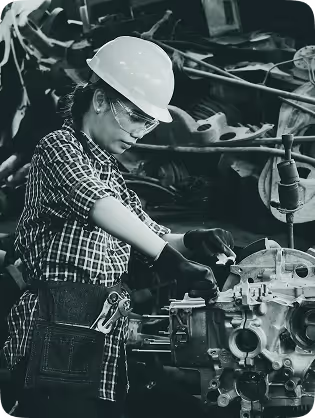 Woman wearing a hard hat and safety glasses working on an engine or mechanical component with gloves in an industrial setting.