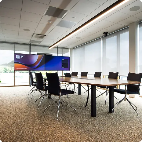 Moderne vergaderruimte met een lange tafel, acht zwarte bureaustoelen en twee grote beeldschermen aan de muur.