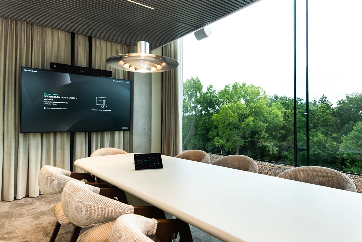 Moderne vergaderzaal met een witte tafel, zes stoffen stoelen, een plafondlamp, een groot scherm aan de muur, en een raam met uitzicht op groene bomen.
