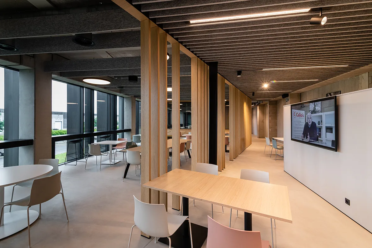 Moderne gemeenschappelijke ruimte met houten scheidingswanden, tafels, stoelen en een grote tv-scherm aan de muur.