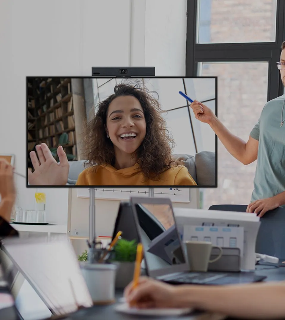 Een man geeft een presentatie aan een groep collega's in een vergaderruimte, terwijl een vrouw op de tv-meeting vrolijk uitzwaait.