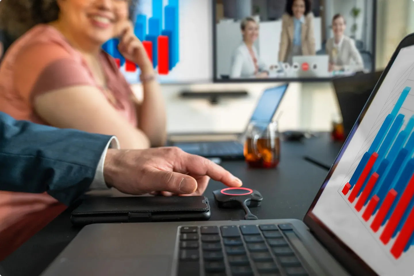 Hand die een draadloze presentatieknop indrukt, met laptops en grafieken zichtbaar tijdens een vergadering.