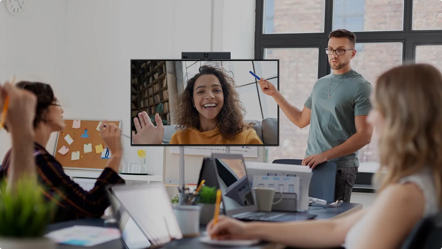 Een man geeft een presentatie aan een groep collega's in een vergaderruimte, terwijl een vrouw op de tv-meeting vrolijk uitzwaait.