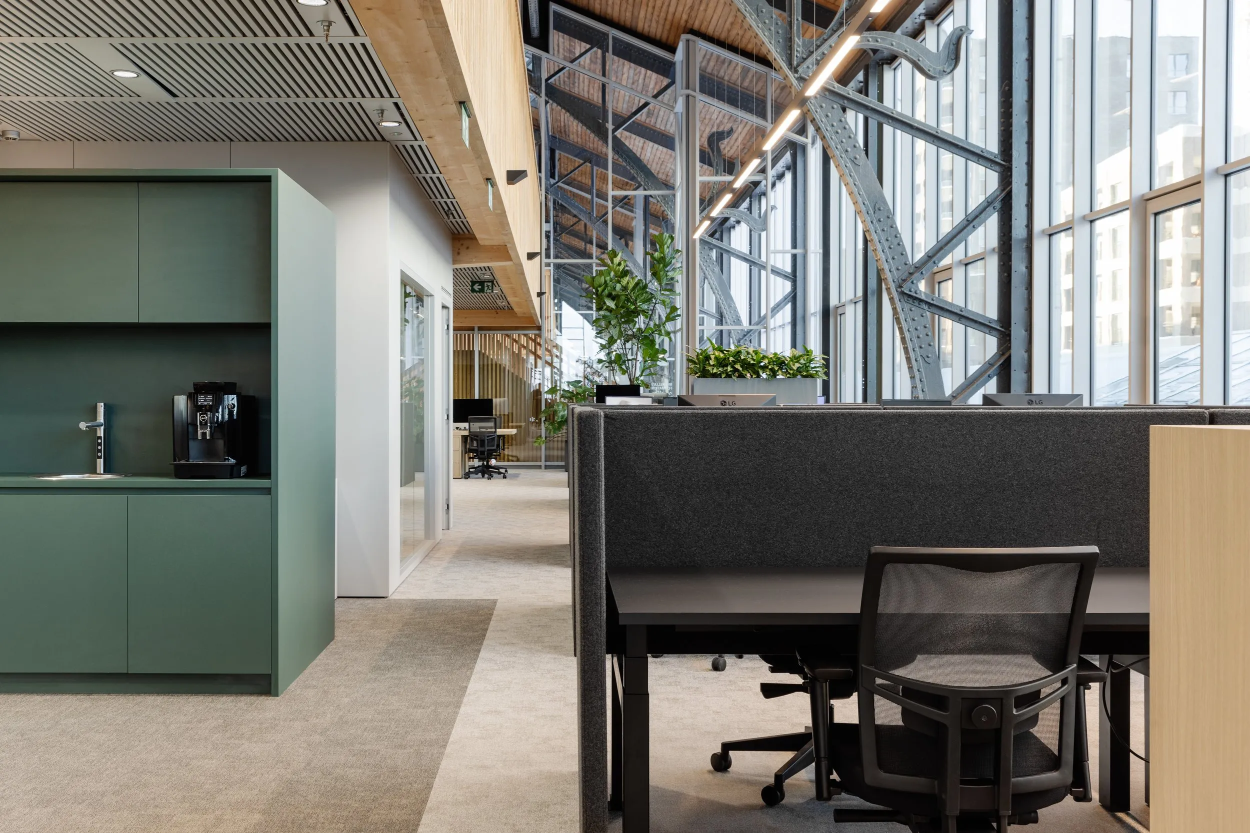 Modern kantoorruimte met groene kasten, koffiezetapparaat, bureaus met geluiddempende scheidingswanden en grote ramen met industrieel staalframe.