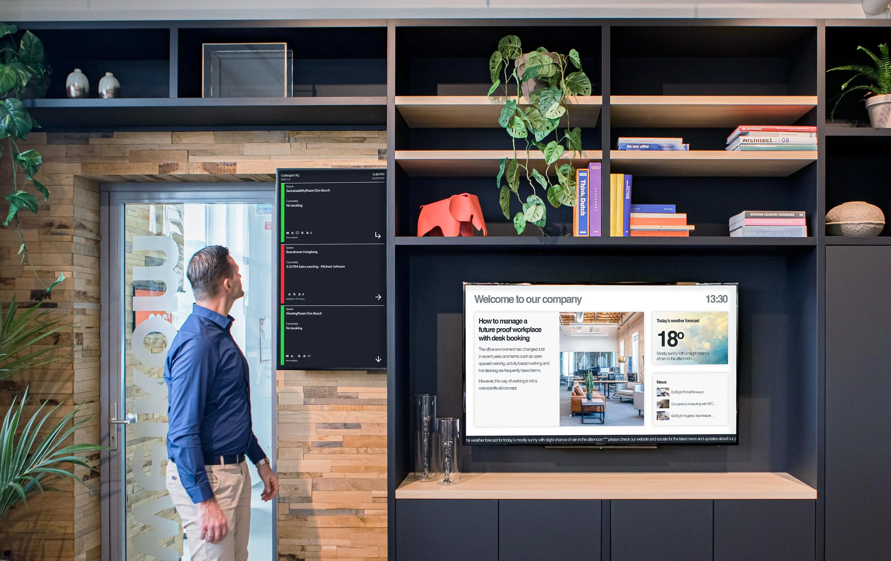 Man kijkt naar digitale schermen met vergaderinformatie en een welkomscherm in een moderne kantoorruimte met houten wand en planten.