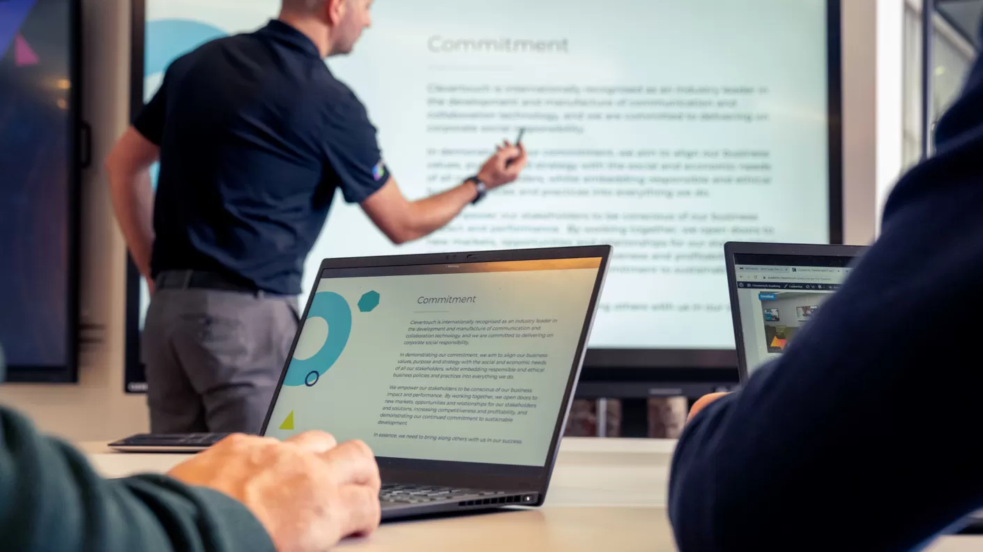 Een man geeft een presentatie in een vergaderruimte terwijl twee mensen met laptops naar het scherm kijken.