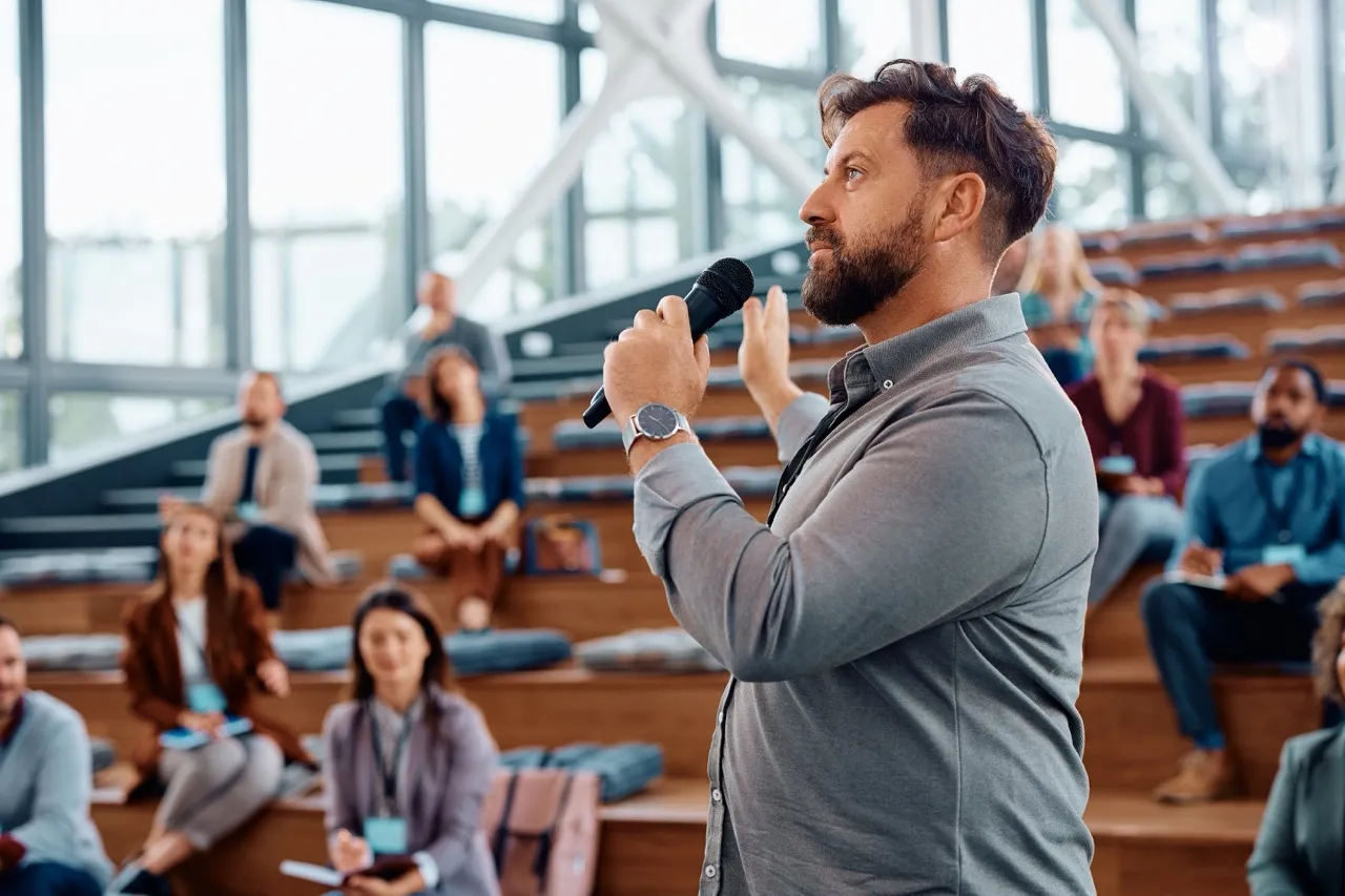 Man met microfoon die spreekt voor een groep volwassenen die op houten tribunes zitten in een moderne, lichte zaal.