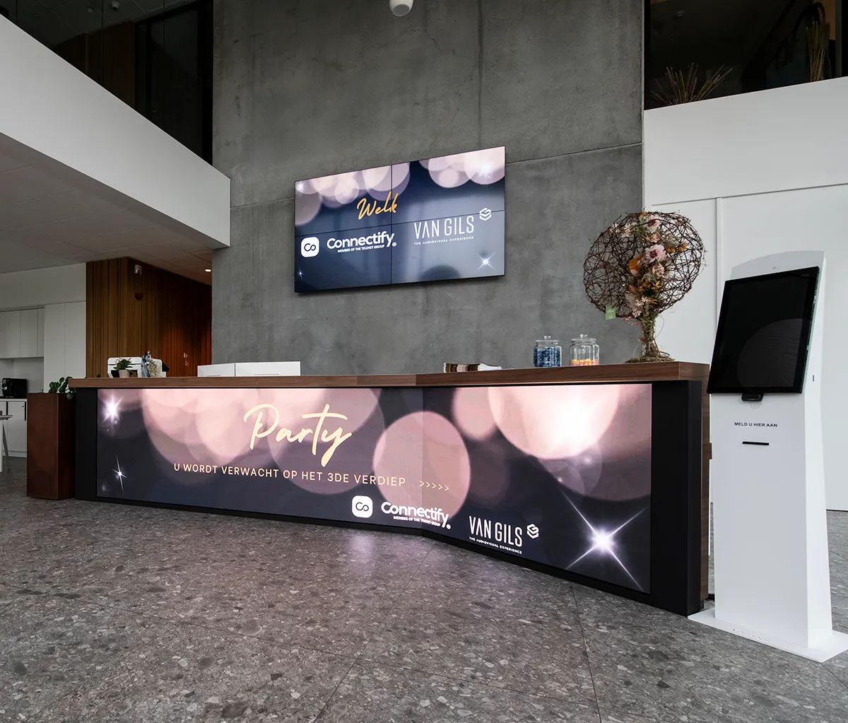 Moderne receptiebalie met digitale schermen die een feest aankondigen op de derde verdieping, en een witte digitale aanmeldzuil rechts.