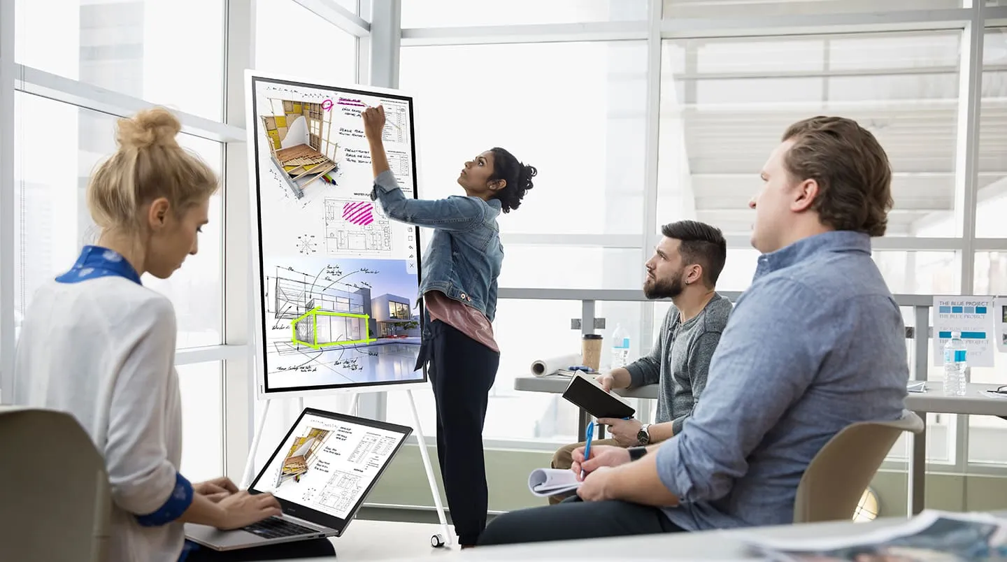 Vrouw presenteert bouwplannen op een digitaal bord terwijl drie collega's luisteren in een training room.
