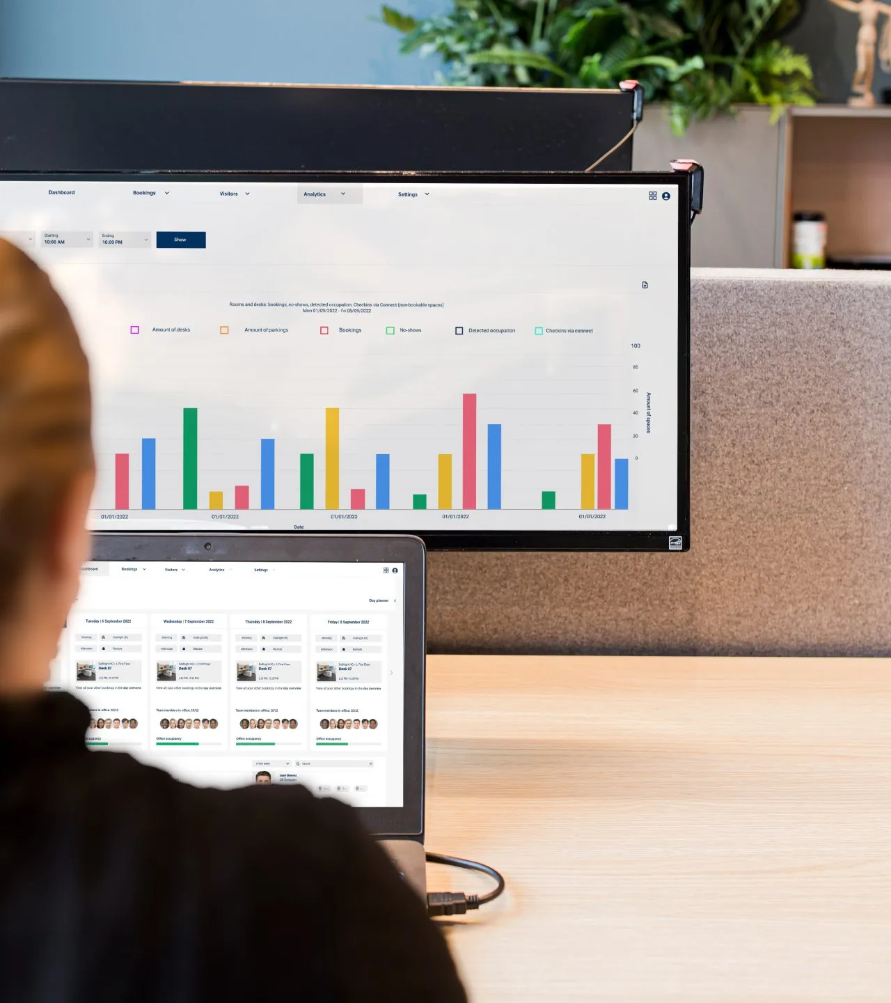 Persoon aan bureau die een laptop gebruikt met grafieken en data-analyses zichtbaar op het laptopscherm en een groter scherm erachter.