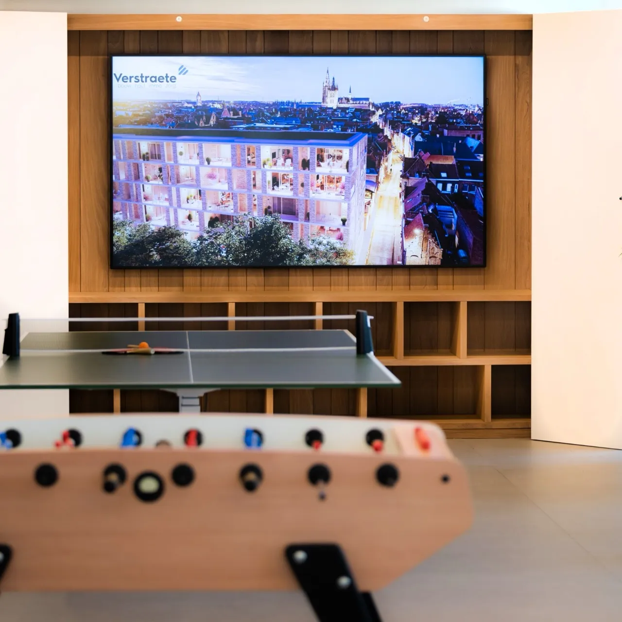 Speelruimte met tafeltennistafel en tafelvoetbalspel, grote scherm aan houten wand toont stadsbeeld met logo Verstraete.
