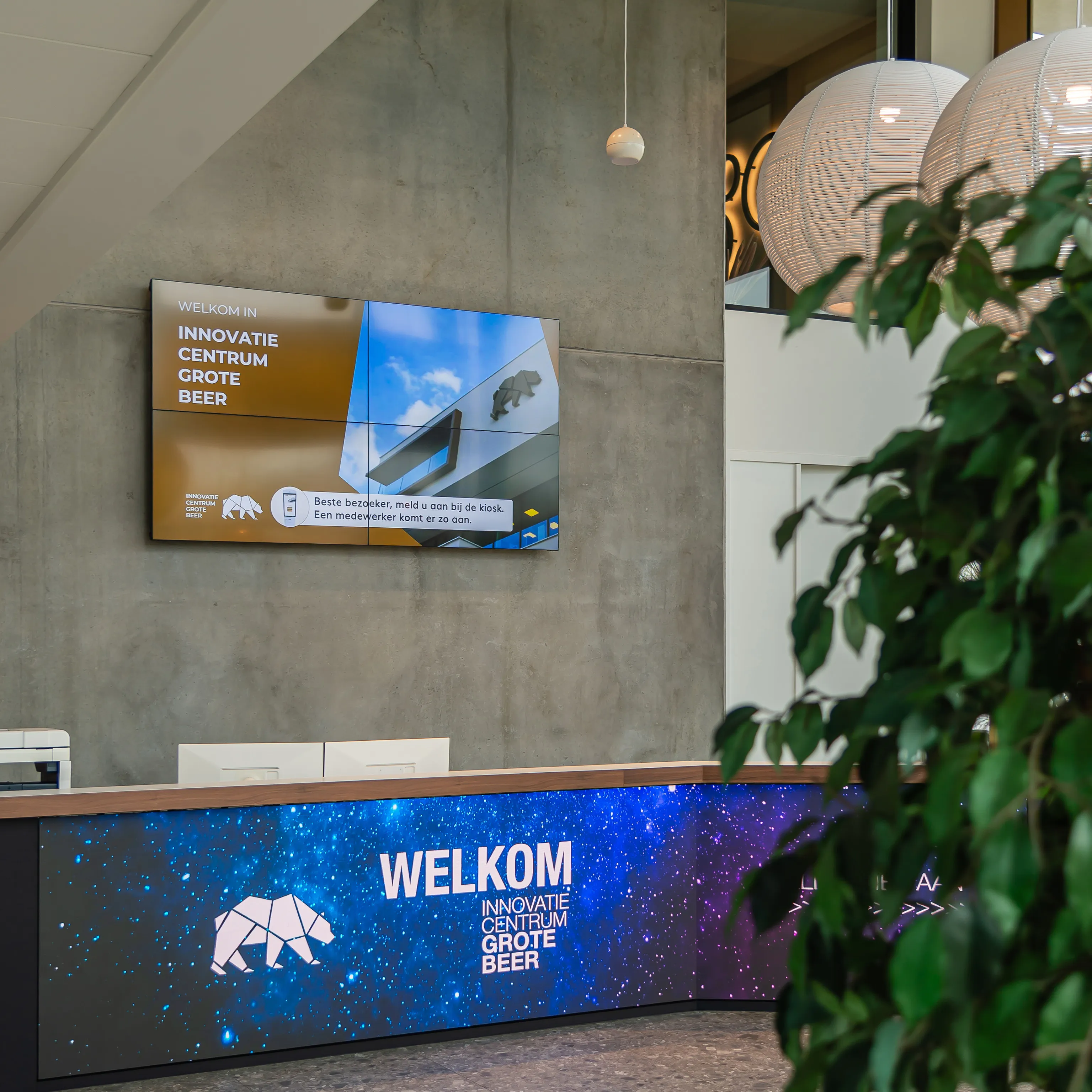Ontvangstbalie in Innovatie Centrum Grote Beer met digitaal bord en plant op de voorgrond.