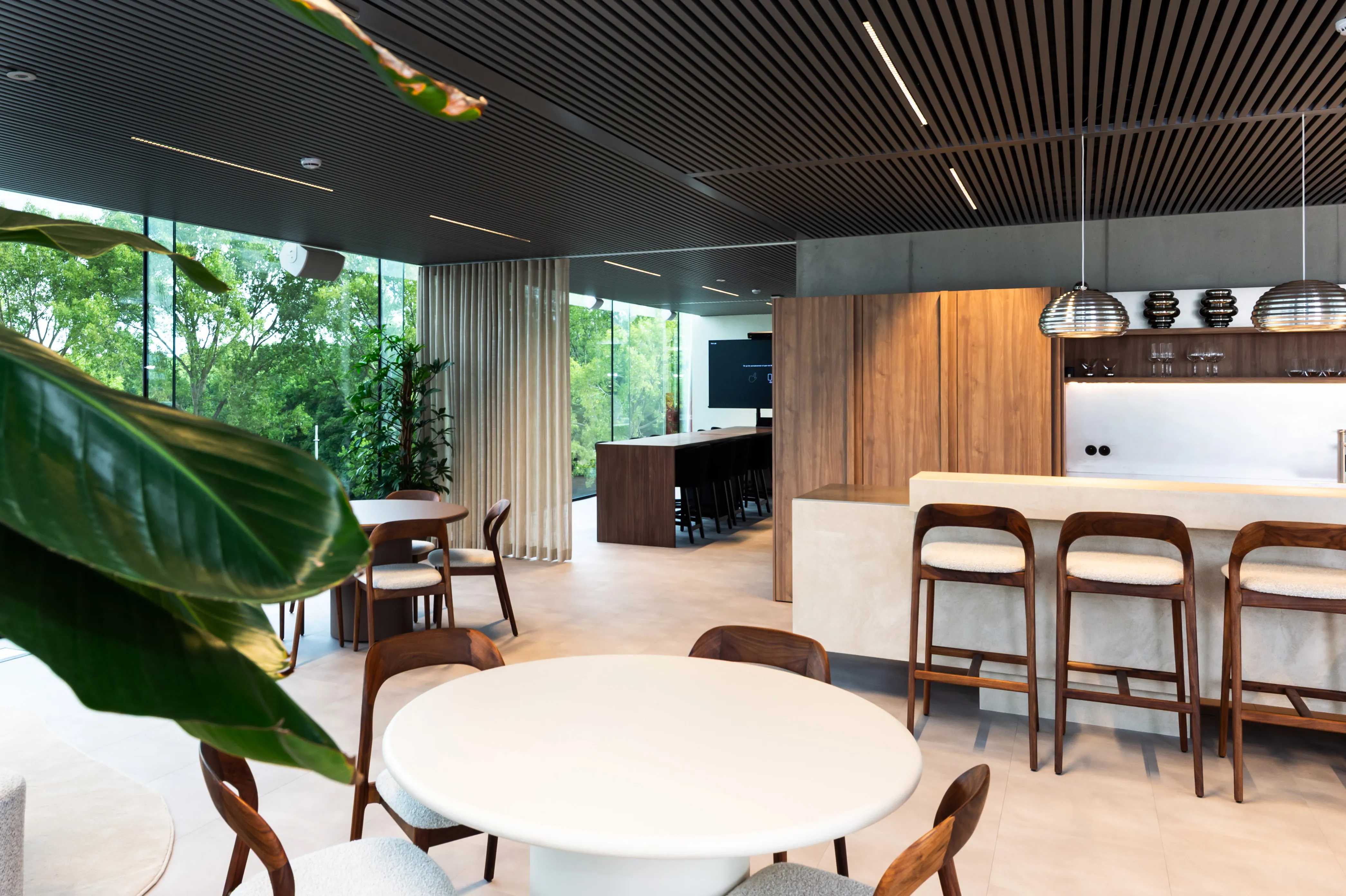 Modern kantoorruimte met ronde tafels, houten stoelen en een bar met barkrukken, omringd door glazen wanden en groene planten.