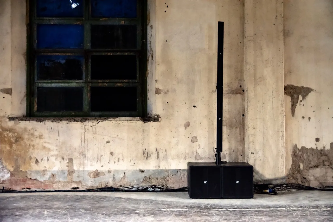 Zwarte line array luidsprekers geplaatst op een betonnen vloer tegen een verweerde muur met een raam.