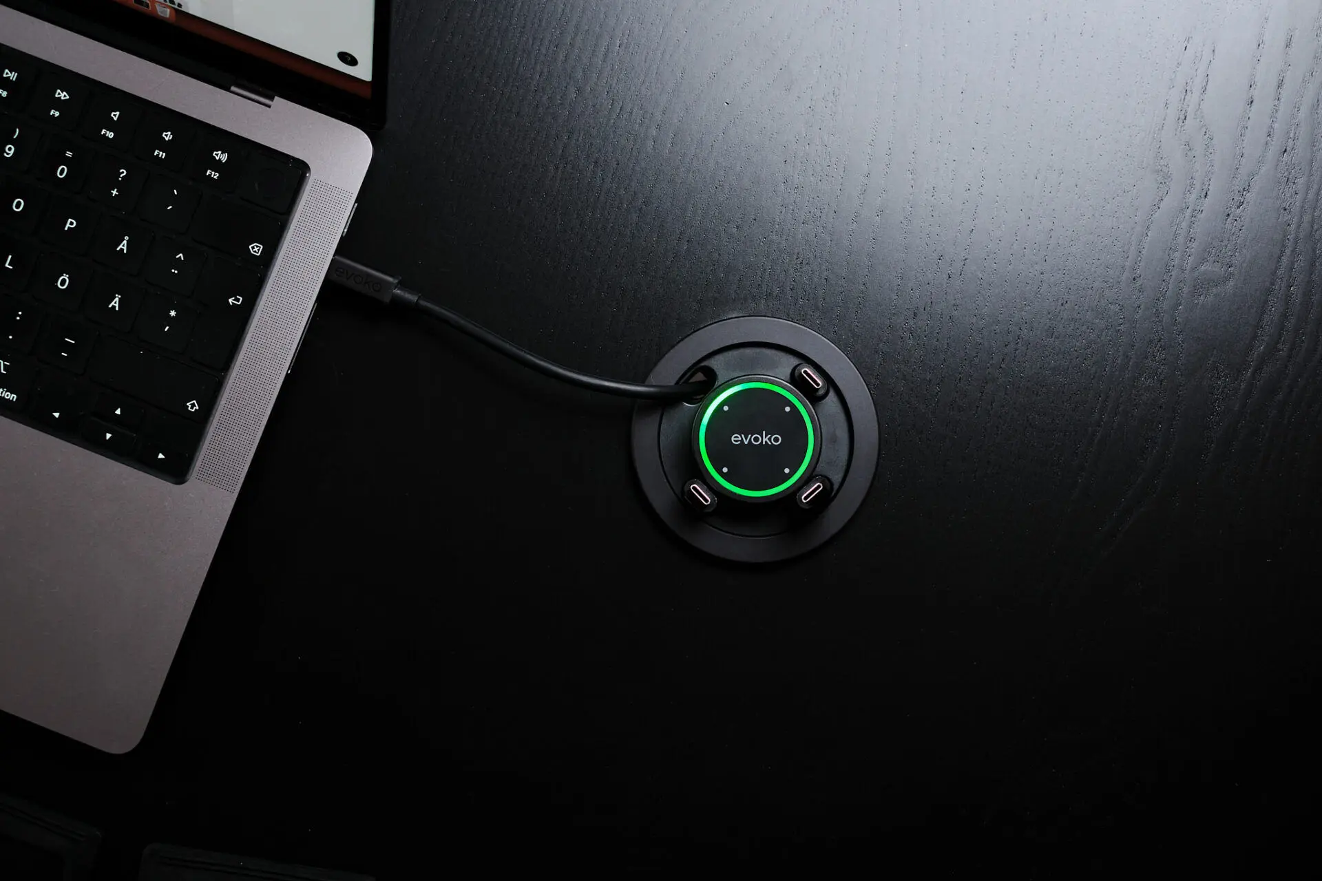 Een zwarte Evoko-kabelverdeeladapter met een groene ring verbonden met een laptop op een zwart houten bureau.