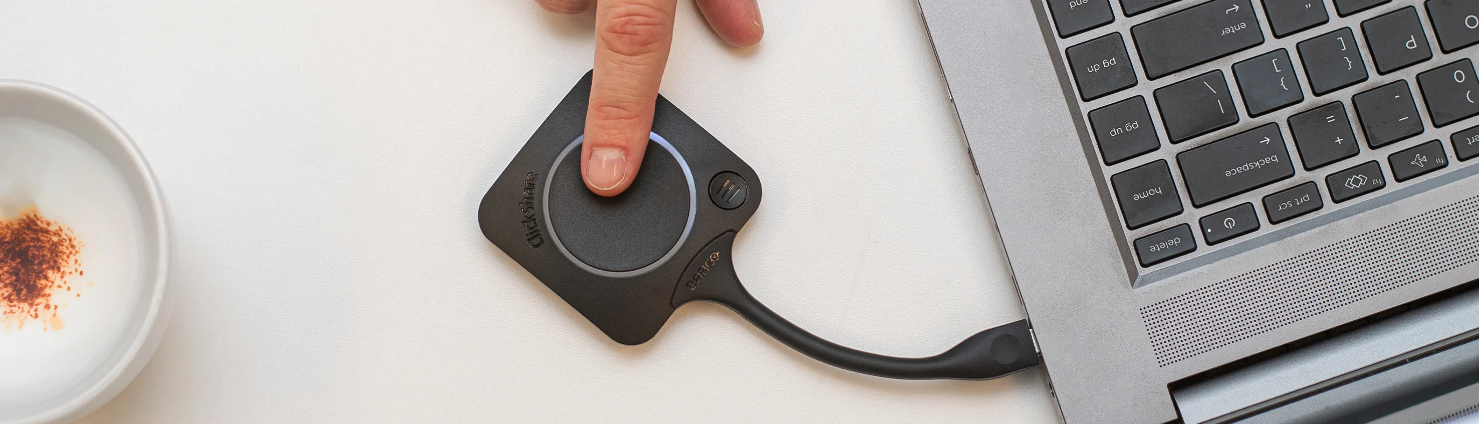 Vinger die op een zwarte Barco ClickShare draadloze presentatieknoop drukt, aangesloten op een laptop naast een kop koffie.