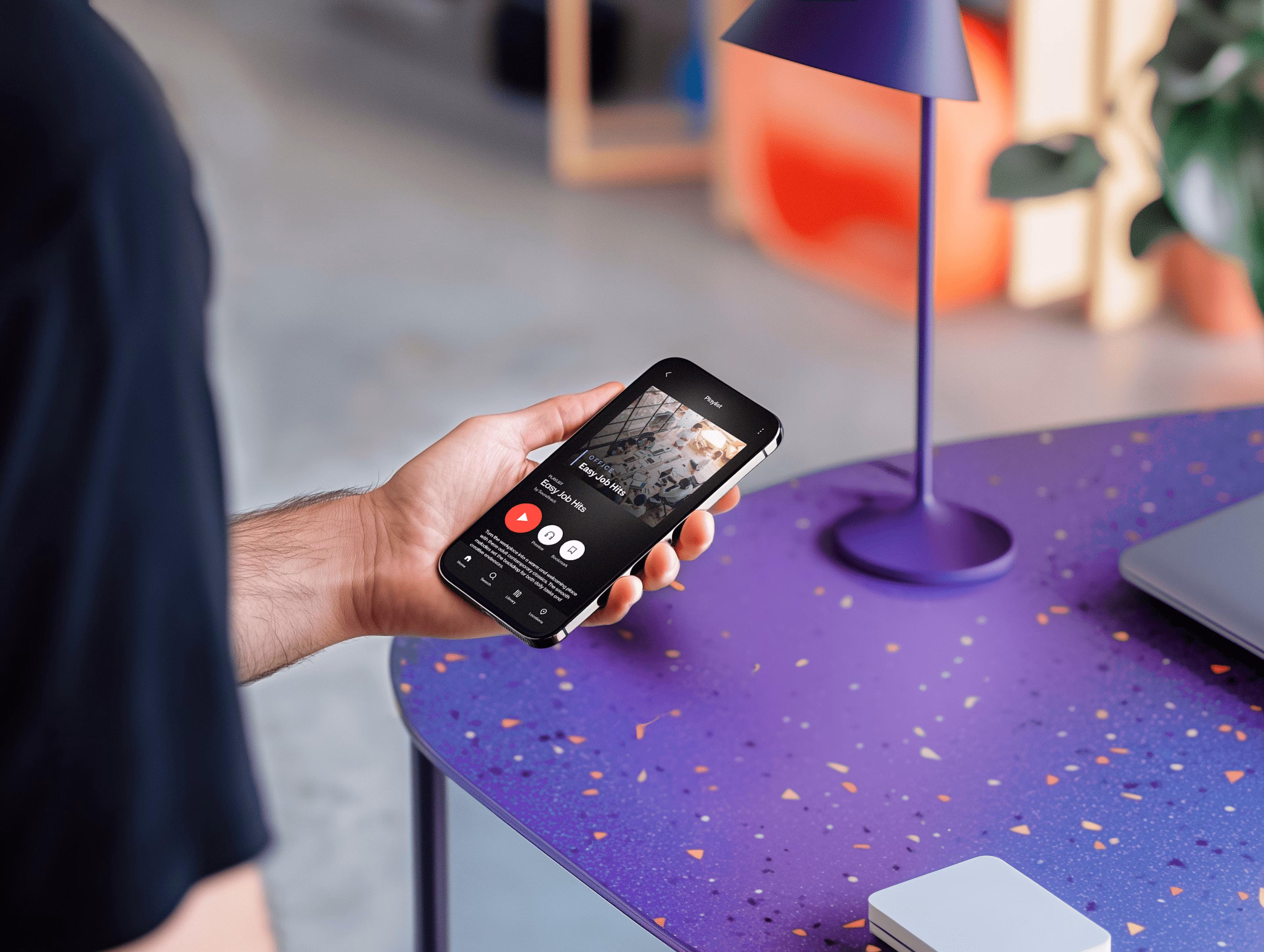 Iemand houdt een smartphone vast met een muziekapp die een afspeellijst 'Easy Job Hits' toont, naast een paars bureau en een paarse lamp.