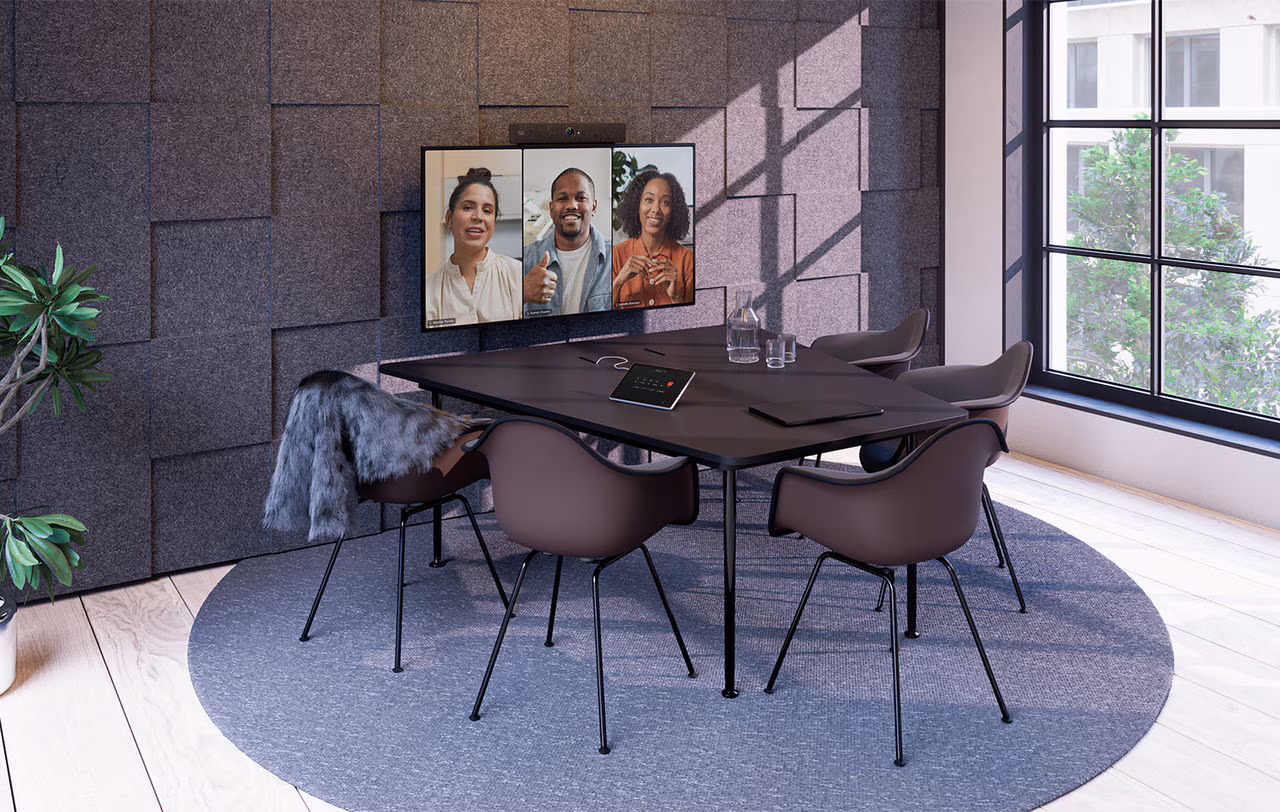 Kleine vergaderruimte met een vierkante tafel, vier stoelen, een grijs vloerkleed en een scherm aan de muur waarop drie mensen videobellen.