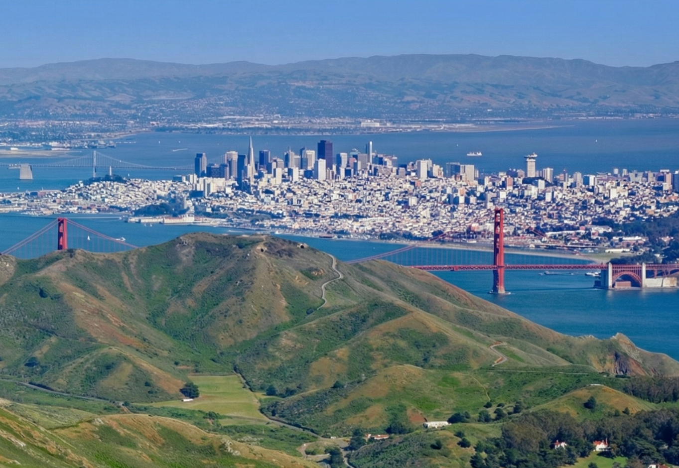 A panoramic aerial view of the rolling green hills of the Marin Headlands with the Golden Gate Bridge spanning across the blue waters of the San Francisco Bay. The San Francisco skyline is visible in the distance under clear blue skies.