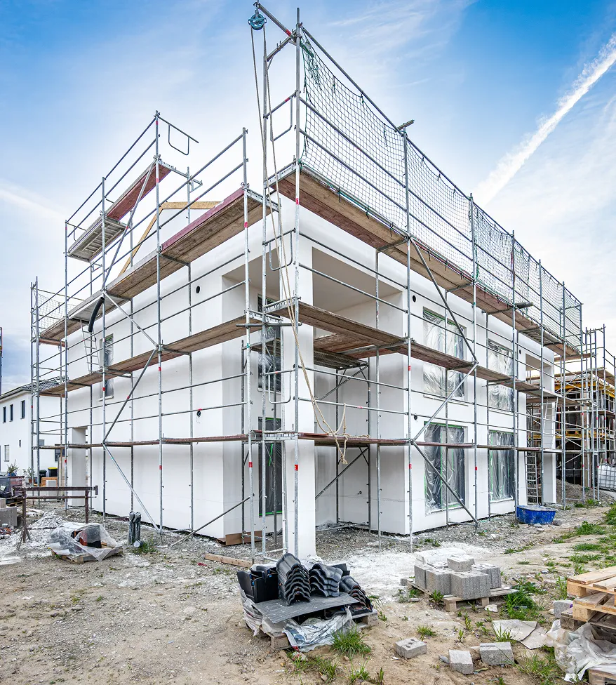 Modernes zweistöckiges Wohnhaus mit weißer Fassade im Rohbau, umgeben von Baugerüsten und Baumaterialien im Vordergrund.