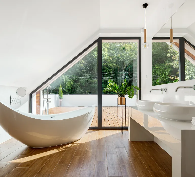 Modernes Badezimmer mit einer freistehenden Badewanne, Doppelwaschbecken und großen Fenstern mit Blick auf einen Balkon und grüne Bäume.