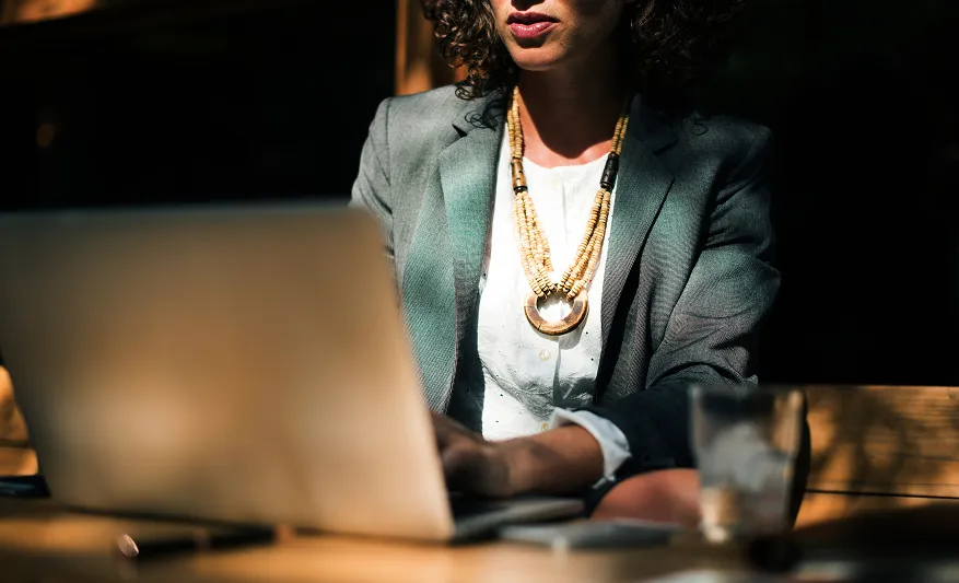 Eine Frau im grauen Blazer sitzt bei Tageslicht an einem Tisch und schaut auf einen Laptop.