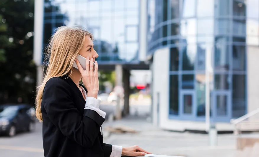 Eine Frau mit langen blonden Haaren spricht draußen am Handy vor modernen Glasgebäuden.