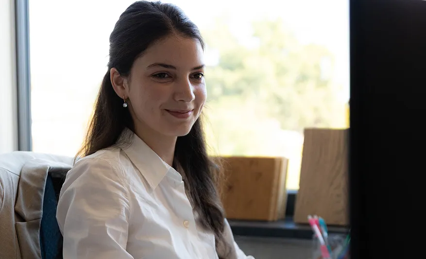 Junge Frau mit weißen Hemd sitzt am Schreibtisch und lächelt, während sie auf einen Monitor blickt.