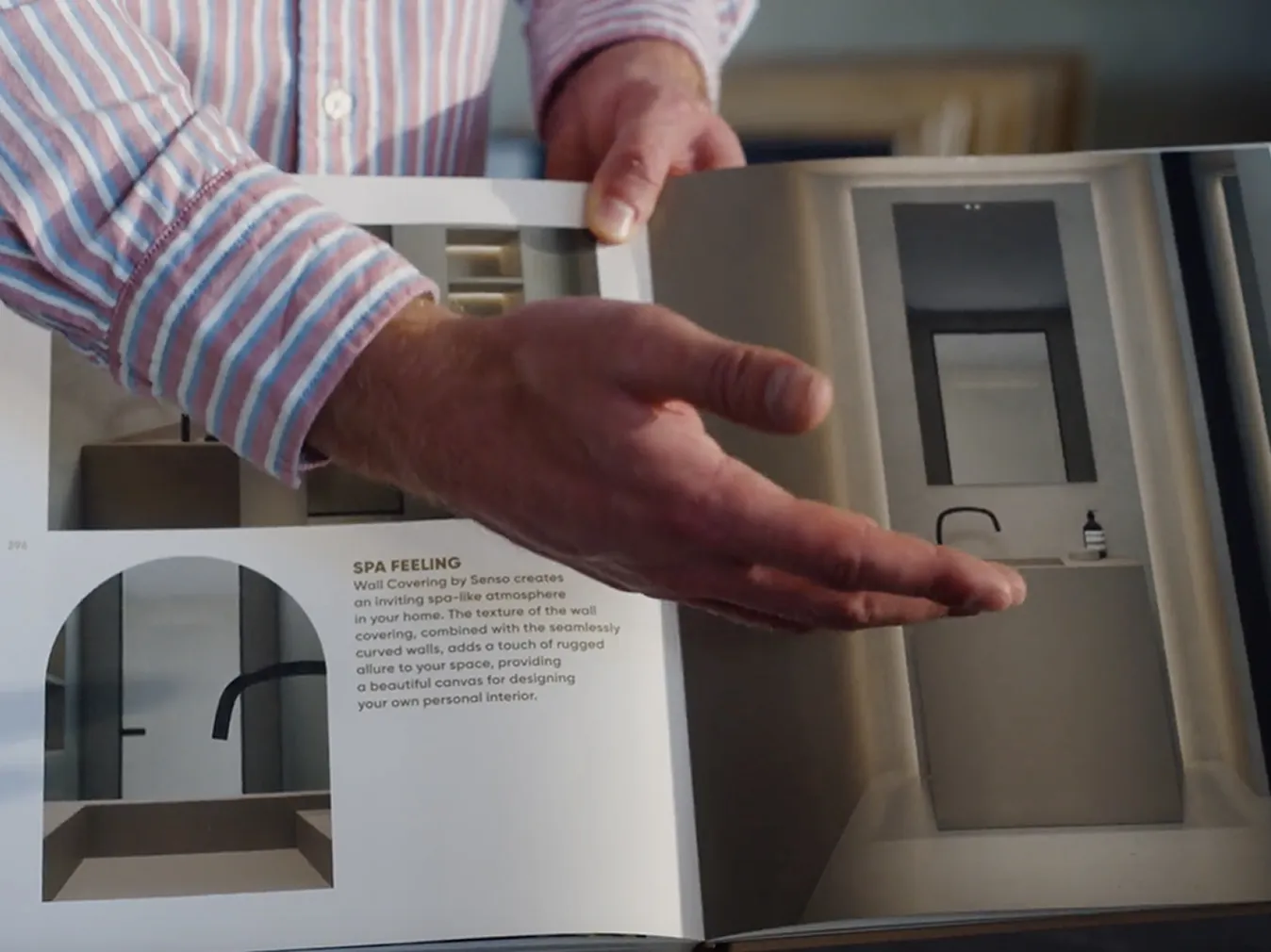 Eine Hand hält ein geöffnetes Buch mit Bildern und Text über ein modernes Badezimmer mit einem Waschbecken, Spiegel und minimalistischer Einrichtung.