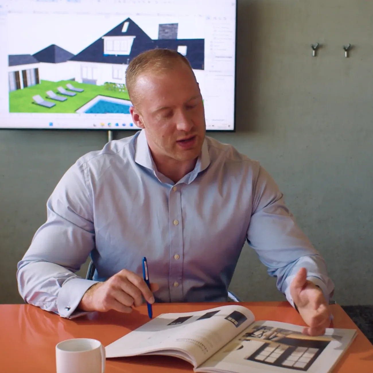 Mann in Hemd erklärt ein geöffnetes Magazin vor einem Bildschirm mit Architekturzeichnung eines Hauses.