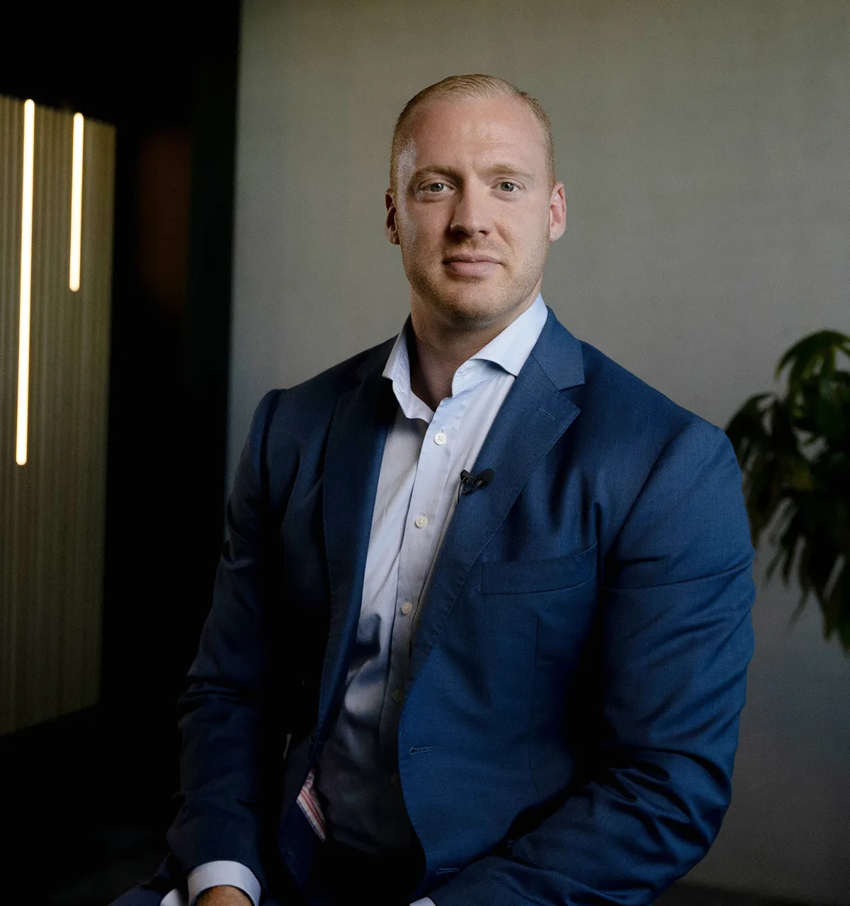 Porträt eines blonden Mannes in einem blauen Anzug und weißem Hemd vor einem neutralen Hintergrund.