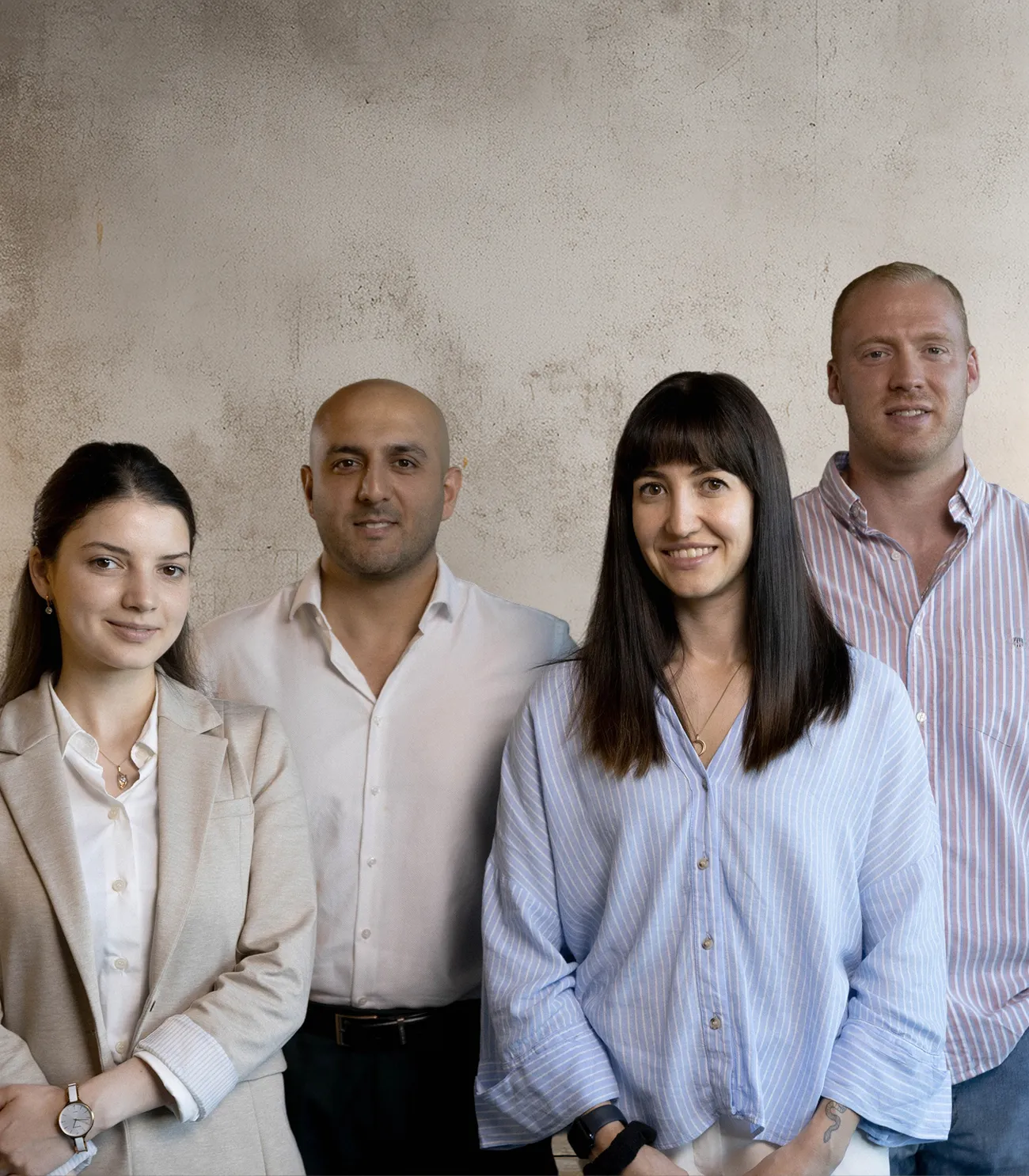 Vier lächelnde Erwachsene, zwei Frauen und zwei Männer, vor einer strukturierten hellen Wand.