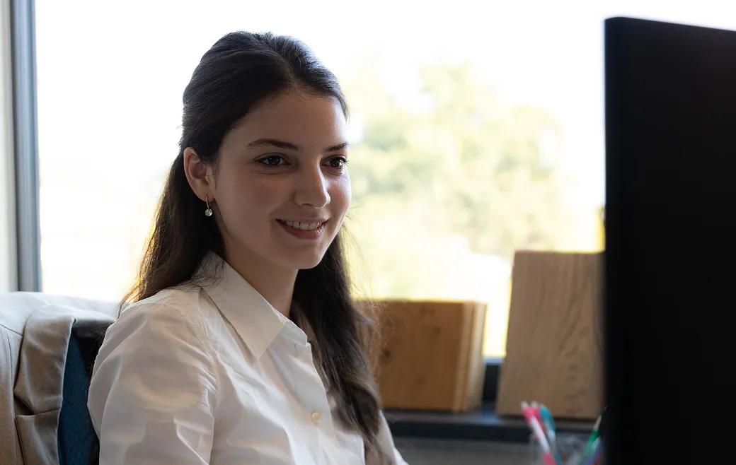 Frau mit langen dunklen Haaren und weißen Hemd lächelt vor einem Computerbildschirm in einem hellen Büro.