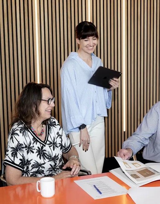 Zwei Frauen, eine sitzt und trägt ein T-Shirt mit Palmenmuster, die andere steht lächelnd mit einem Tablet, im Hintergrund vertikale Holzlamellen.