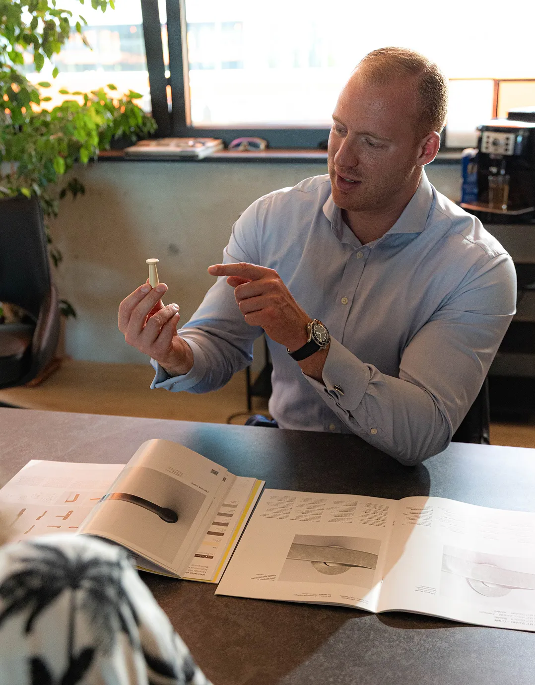 Mann in hellblauem Hemd sitzt an einem Tisch und zeigt auf ein kleines, weißes Objekt, während vor ihm auf dem Tisch mehrere geöffnete Kataloge oder Broschüren liegen.