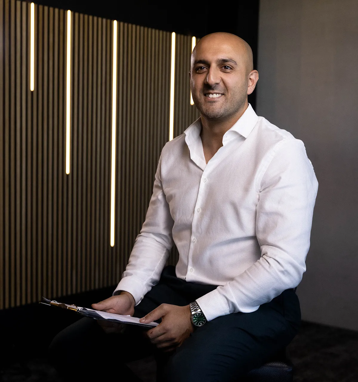 Ein Mann in weißem Hemd sitzt mit einem Klemmbrett in der Hand vor einer Wand mit vertikalen Lichtern.