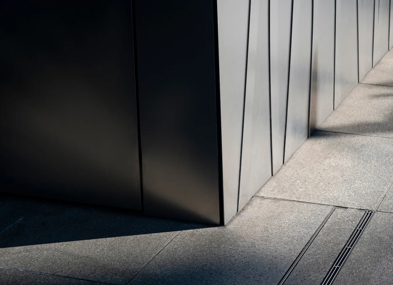 Ecke eines modernen Gebäudes mit grauen Metallplatten und Schatten auf einem hellen Bürgersteig.