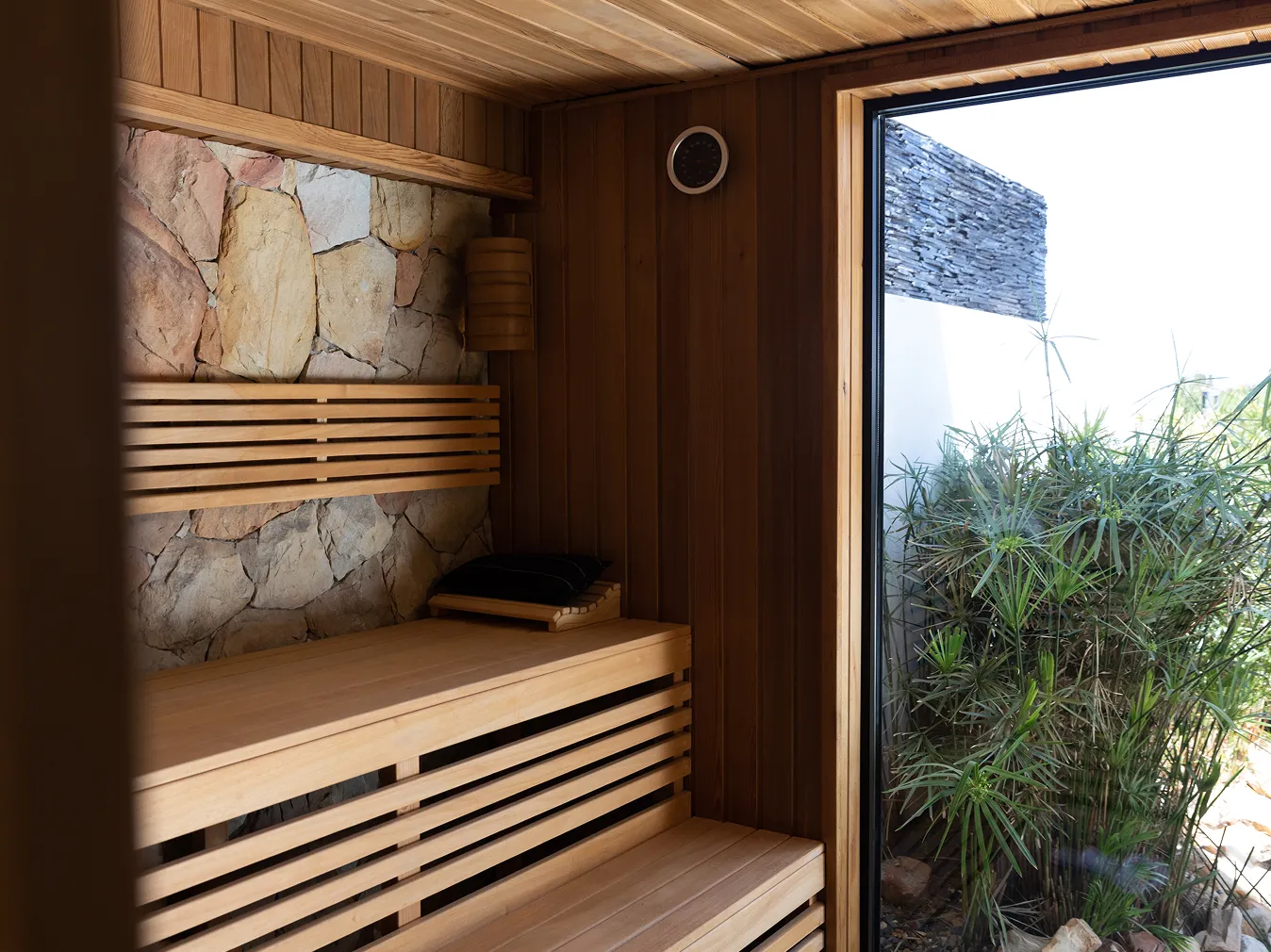 Saunaraum mit Holzverkleidung, Steinwand und großer Glasscheibe mit Blick auf Pflanzen im Freien.