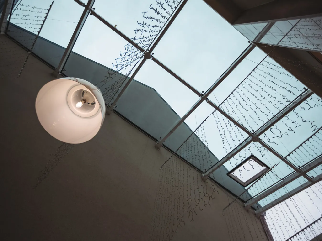 Blick von unten auf ein Glasdach mit hängenden Lichterketten und einer weißen Deckenlampe mit sichtbarer Glühbirne.