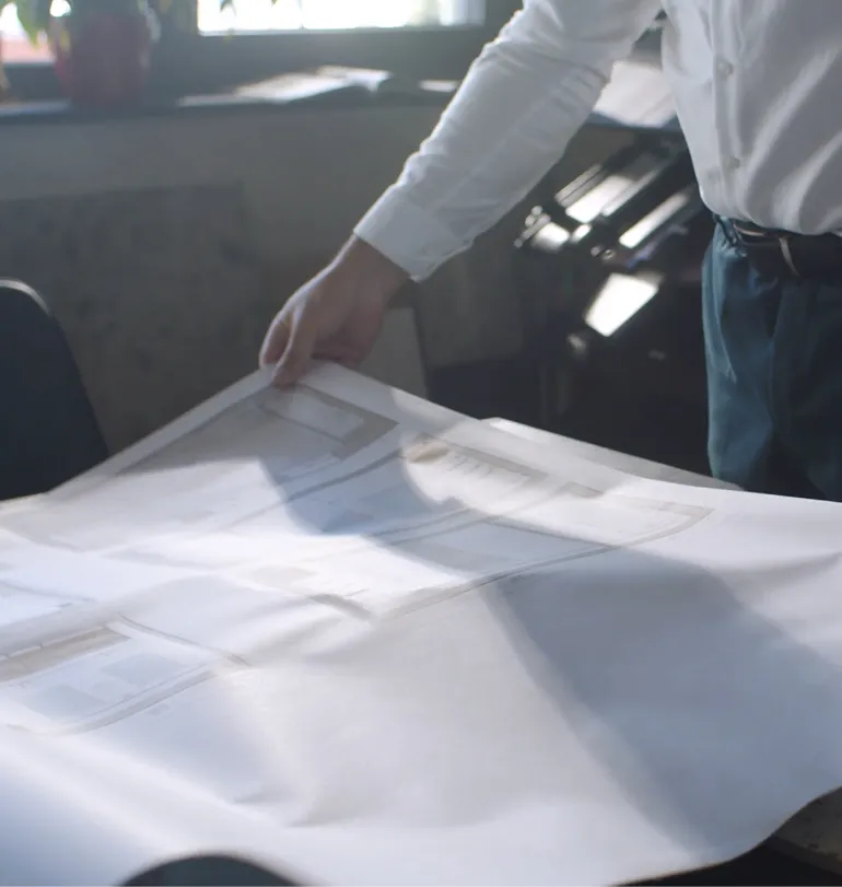 Person im weißen Hemd hält technische Zeichnung oder Bauplan in einem lichtdurchfluteten Raum.