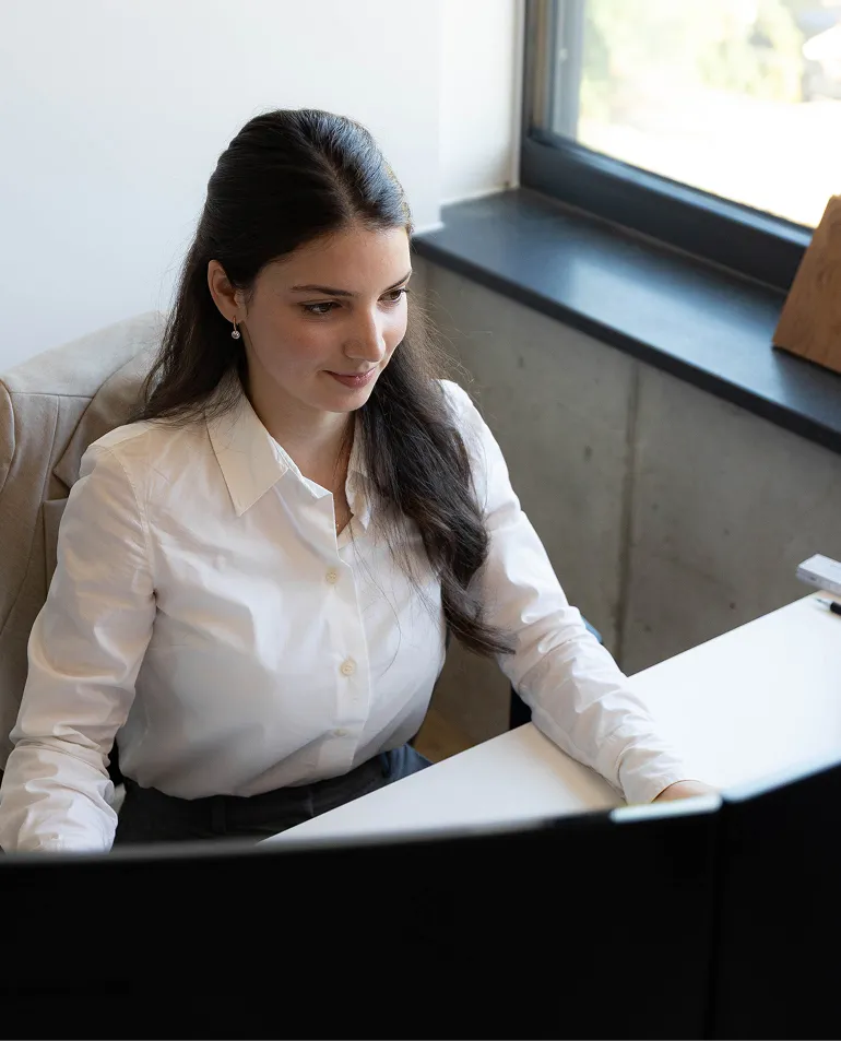 Frau mit weißen Hemd sitzt an einem Schreibtisch und arbeitet an einem Computer vor einem Fenster.