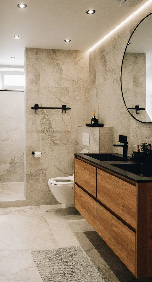 Modernes Badezimmer mit hängendem WC, Holzwaschtisch mit schwarzer Doppelspüle und rundem Spiegel an beigen Steinwänden.