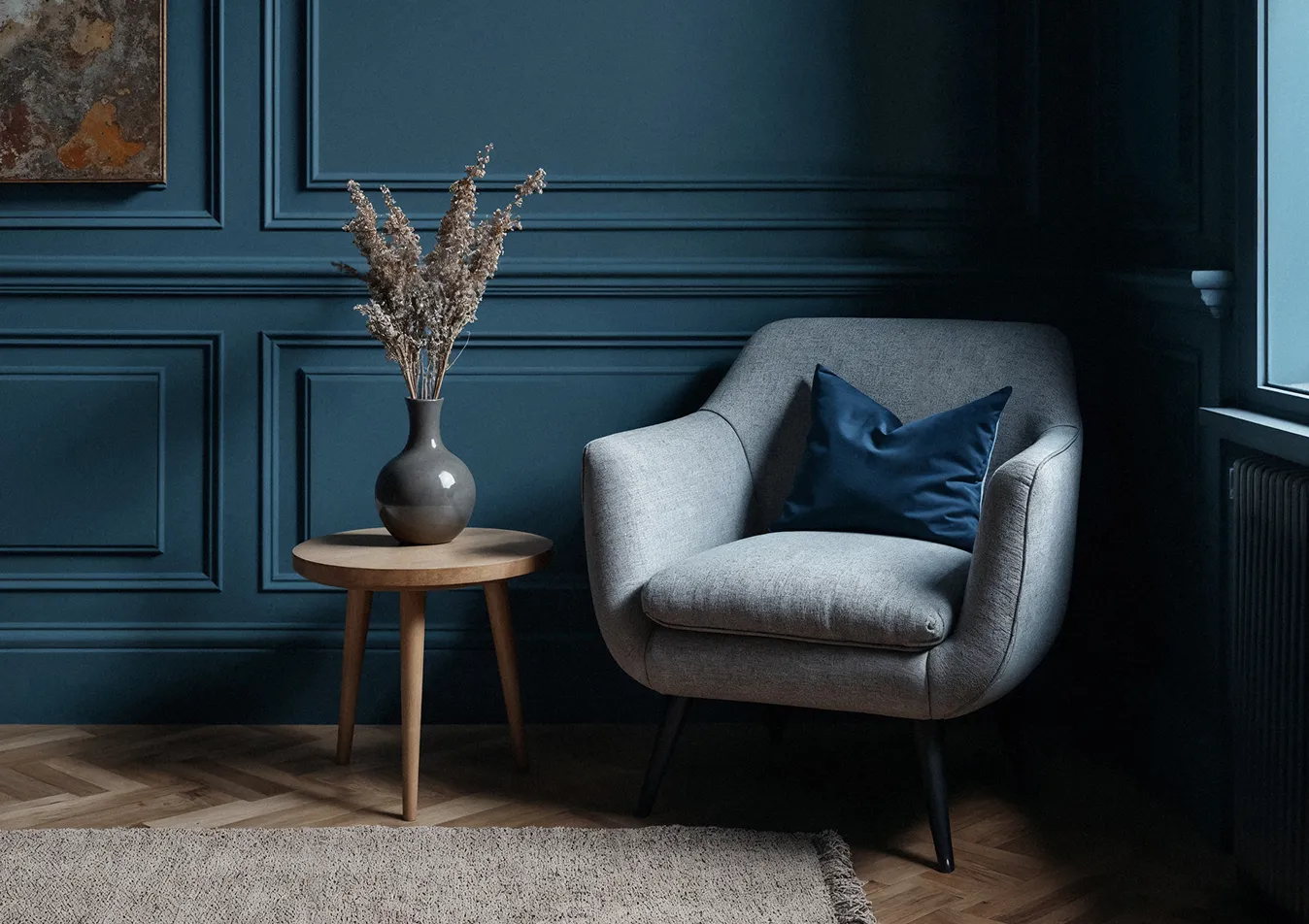 Grauer Sessel mit blauem Kissen neben einem kleinen Holztisch mit einer Vase und getrockneten Blumen vor einer dunkelblauen Wand.