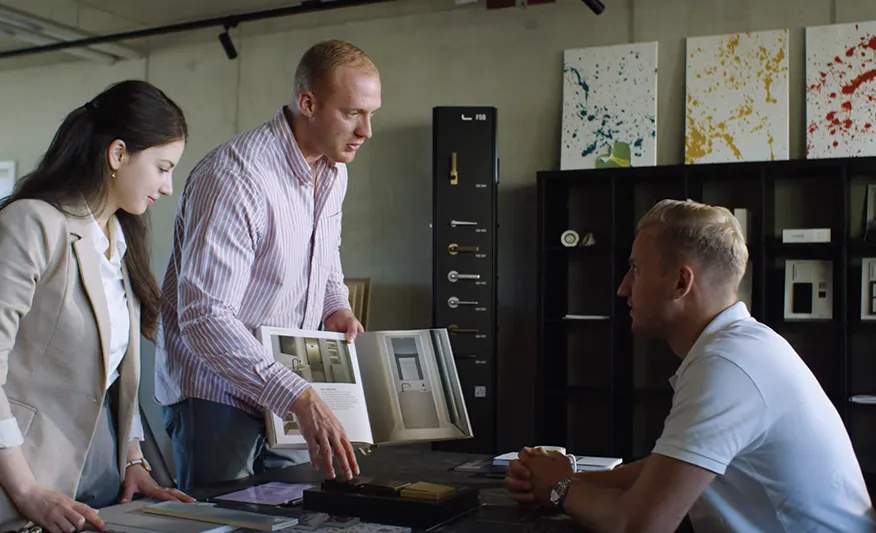 Drei Personen in einem modernen Büro diskutieren über Designmuster, wobei eine Person ein Buch mit Innentür-Designs zeigt.