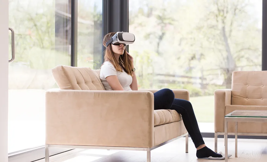 Frau sitzt auf einem beigen Sofa vor großen Fenstern und trägt eine Virtual-Reality-Brille.