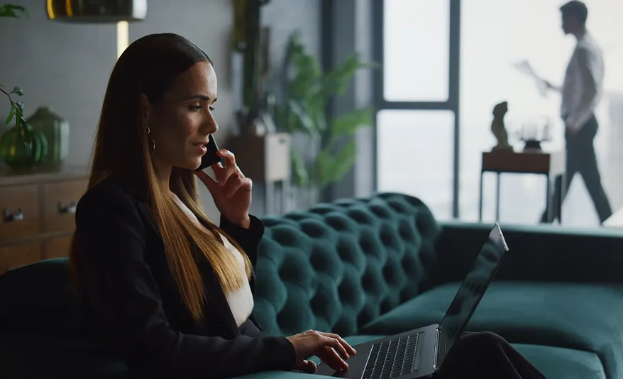 Frau sitzt auf einem dunkelgrünen Samtsofa, telefoniert und arbeitet an einem Laptop in einem modernen Büro.