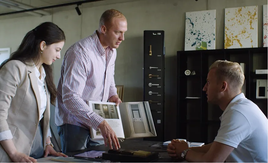 Drei Personen in einem Büro, zwei stehen und zeigen einem sitzenden Mann ein offenes Buch mit Bildern von Türen.